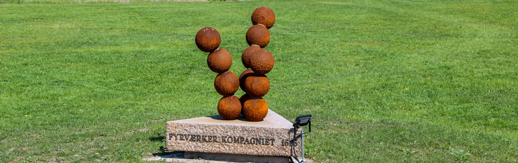 Rustfarvede kanonkugler stablet i tre søjler på granitsokkel