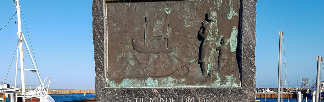 Høj firkantet stentavle med lys skrift og billede af fiskekutter og personer