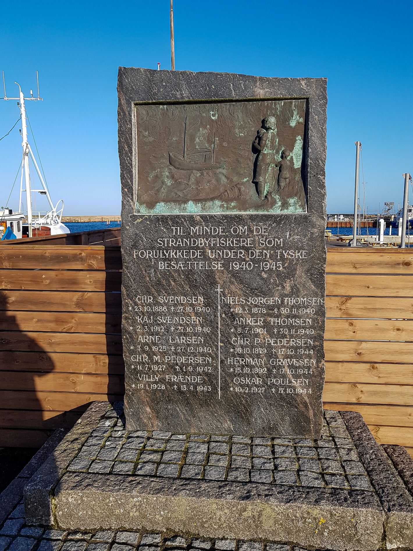 Høj firkantet stentavle med lys skrift og billede af fiskekutter og personer